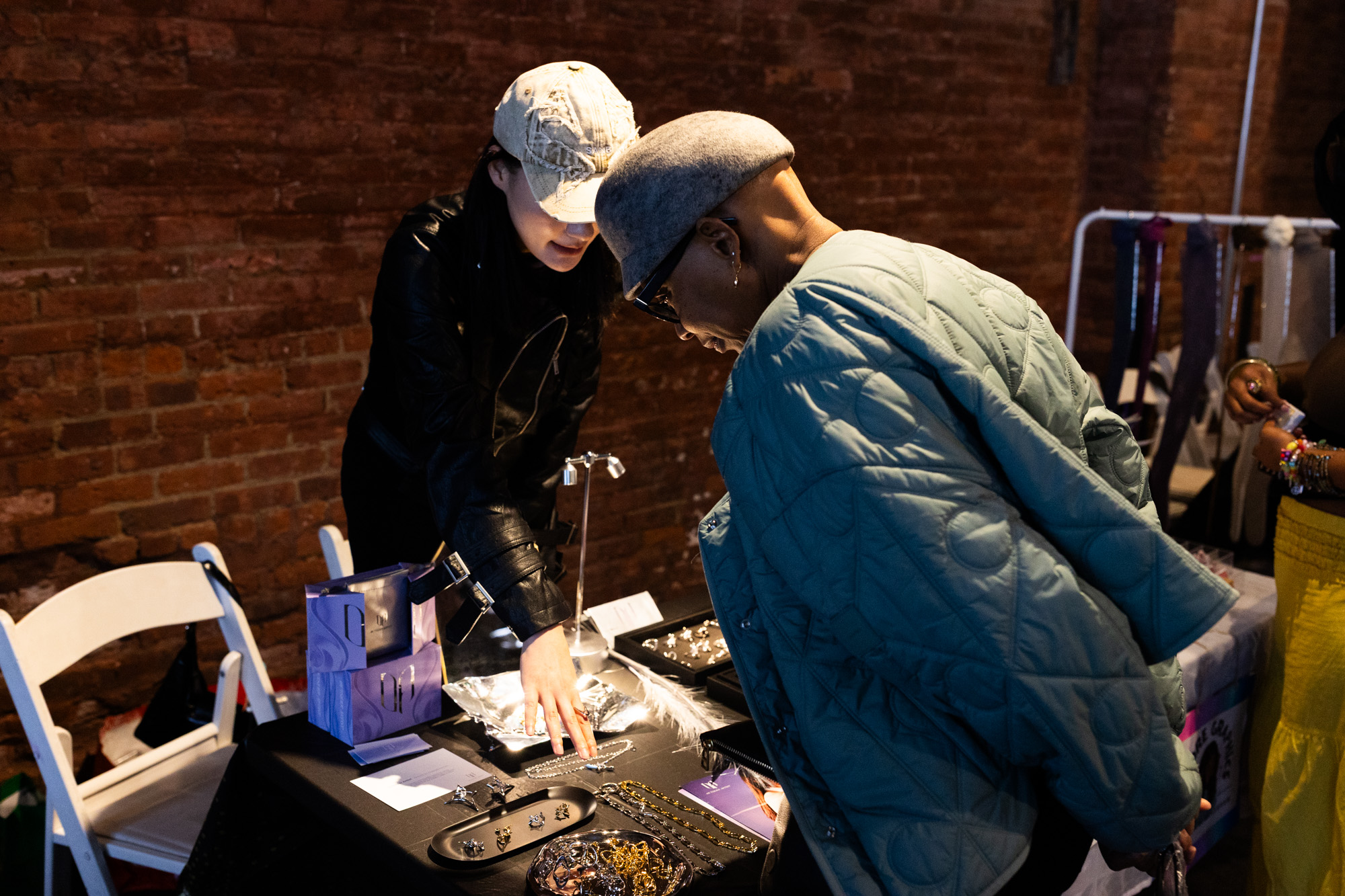 Two attendees leaning in at a jewelry vendor table in warm brick light
